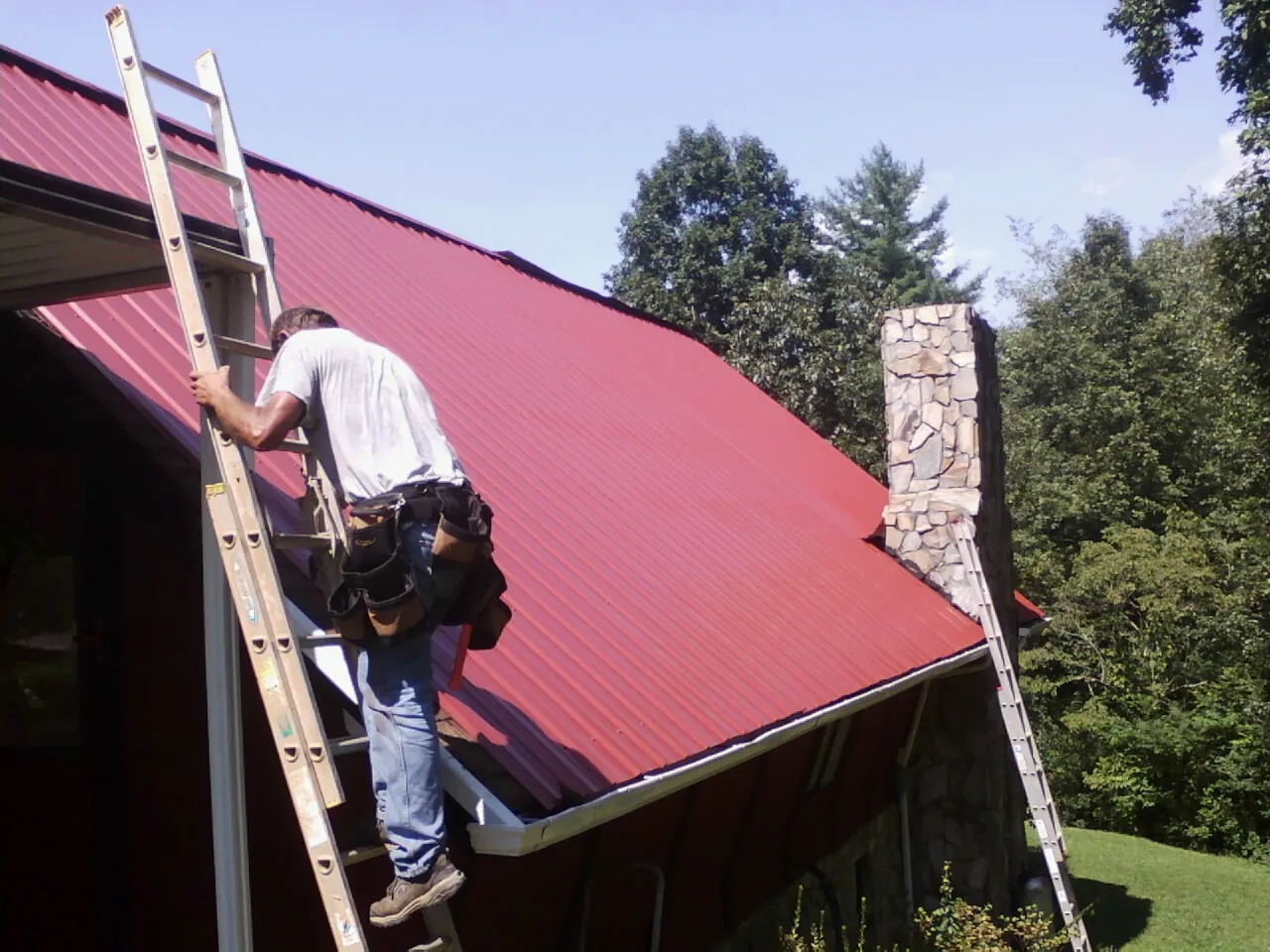 Roofer on a ladder beside a red metal roof and stone chimney — Soffit & Fascia Repair by Roofing Brothers in Eidson